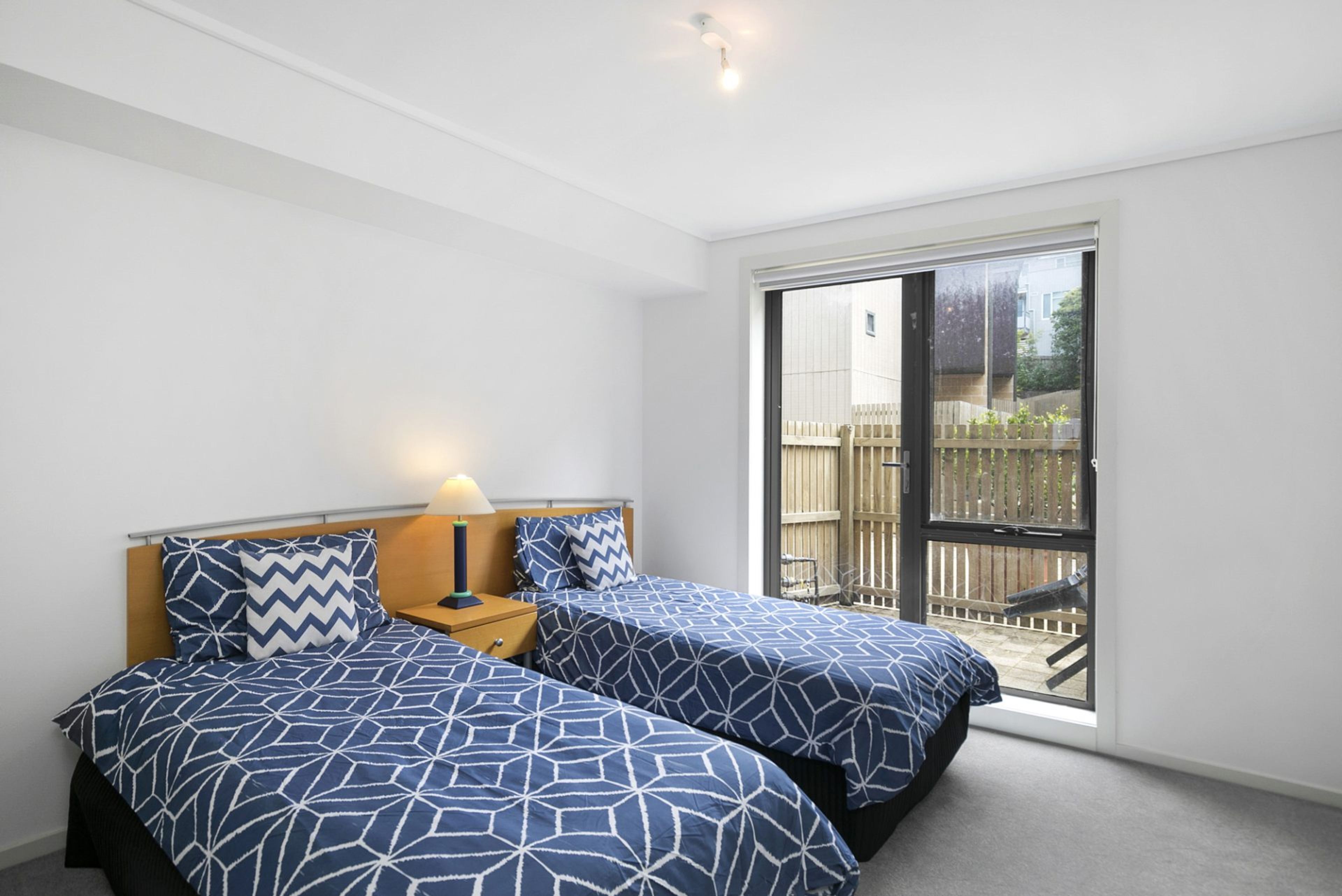 A bedroom with two beds, a nightstand, a lamp, and a sliding glass door leading to an outdoor area.