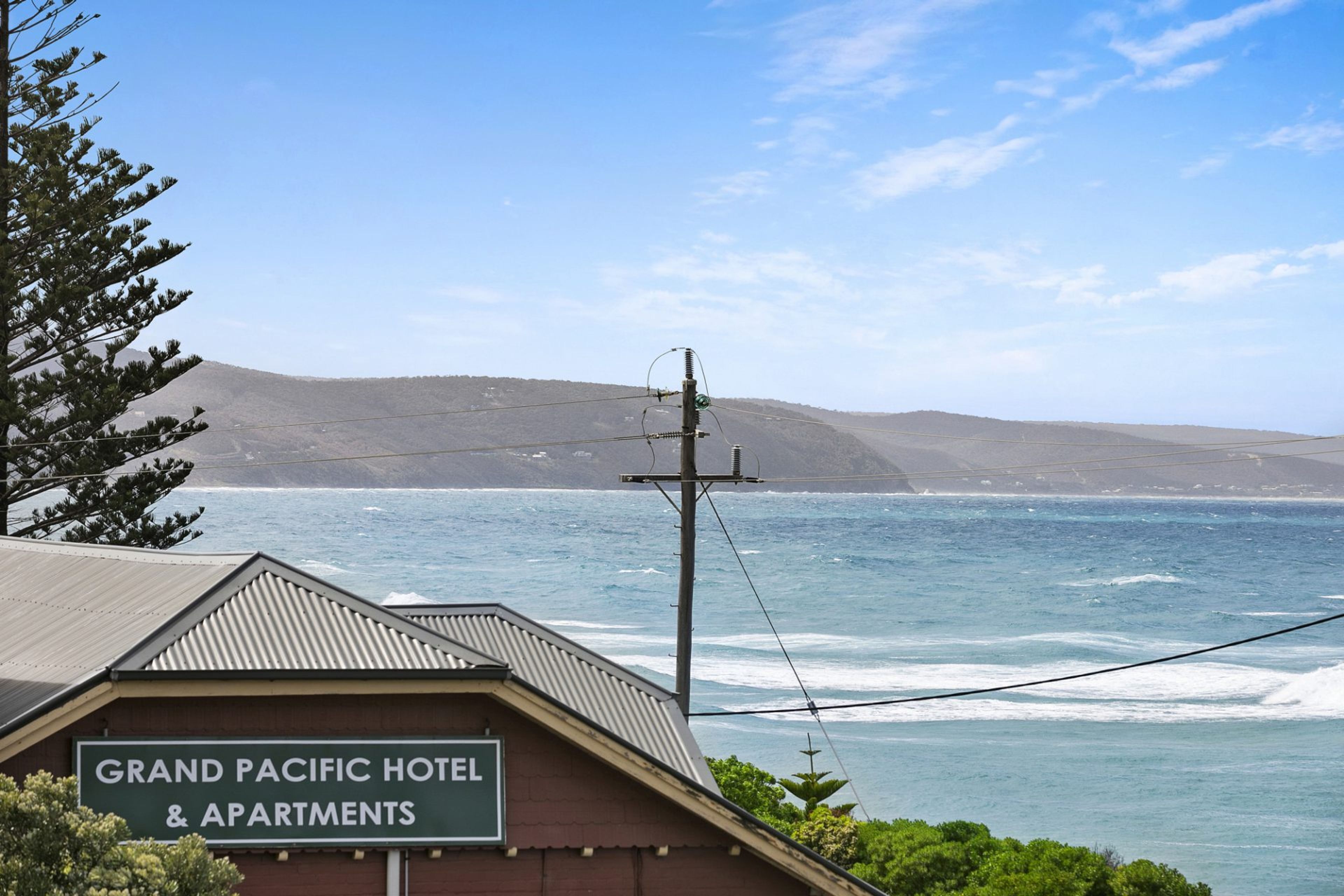 A sign for the Grand Pacific Hotel & Apartments with a view of the ocean and hills in the background.