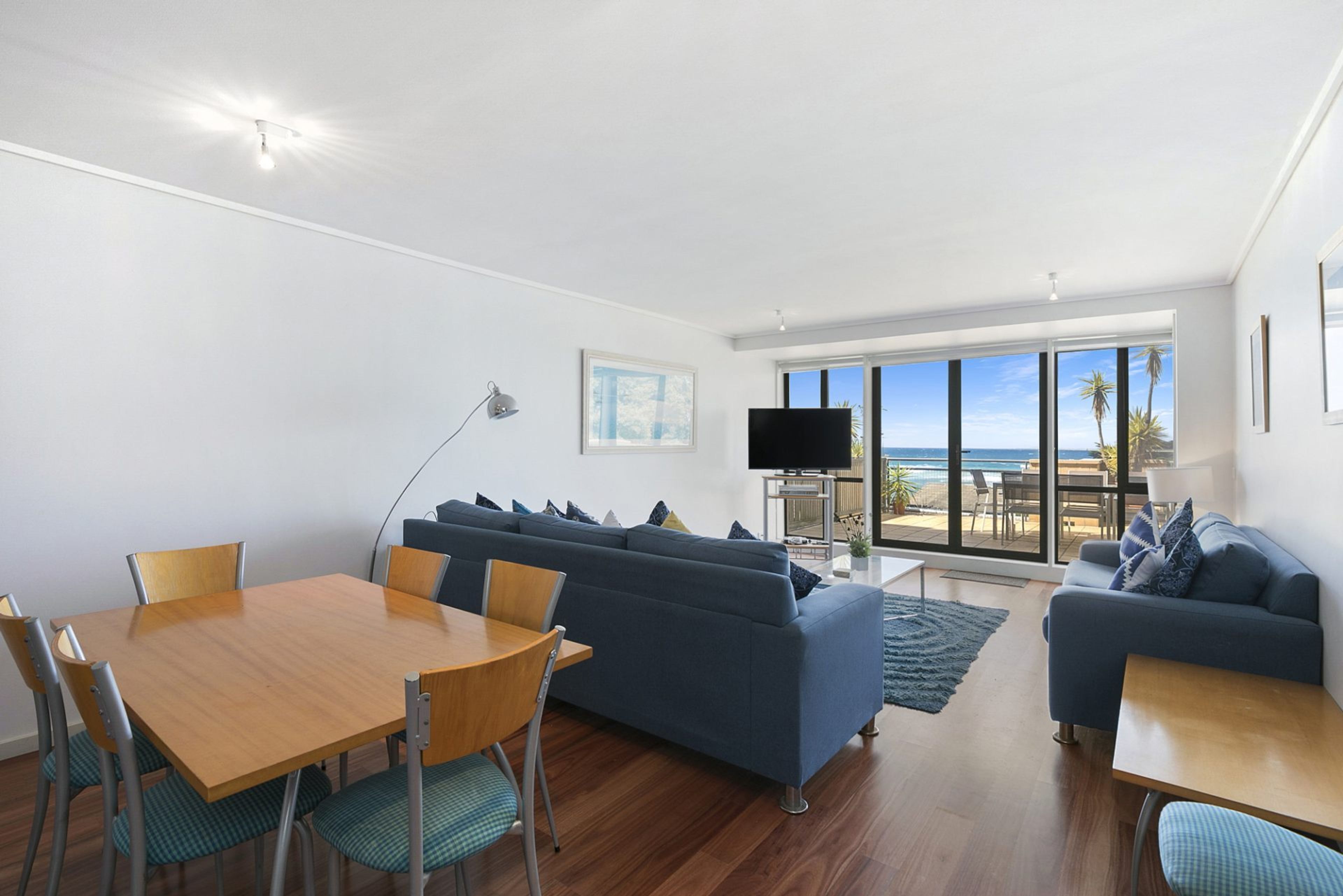 A modern living room with a wooden dining table, blue sofas, and a TV.