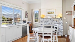Una cocina moderna con armarios blancos, una zona de comedor con sillas blancas, una chimenea de piedra y vistas al exterior.