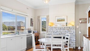 A modern kitchen with white cabinetry, a dining table with chairs, a stone fireplace, and a view through the window. - The White House -Prime Location & Oozing Character (Apollo Bay)