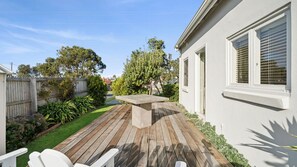 Una terraza de madera con una mesa y sillas, rodeada de un jardín cuidado y un edificio blanco con ventanas.