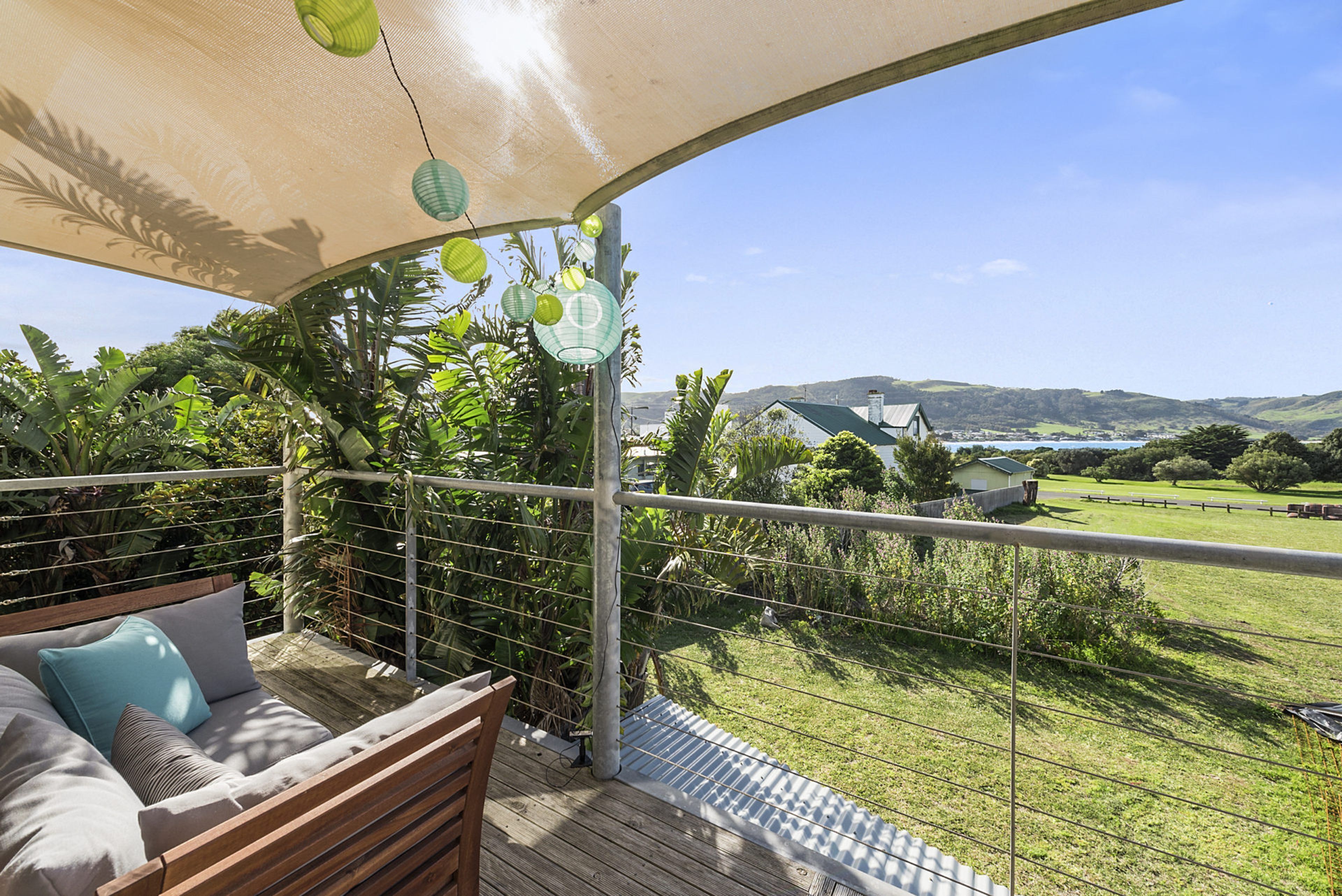 A balcony with a wooden deck, a cushioned seating area, and a view of a green landscape with a building in the distance.