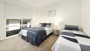 A bedroom with two beds, a window with blinds, a bedside table with a lamp, and a framed picture on the wall.