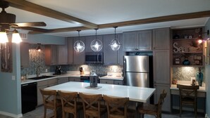 A modern kitchen with a central island, wooden chairs, a stainless steel refrigerator, and pendant lighting.
