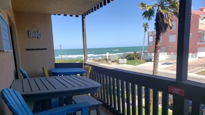 A balcony with blue chairs and a table, overlooking a beach with palm trees and a building in the background.