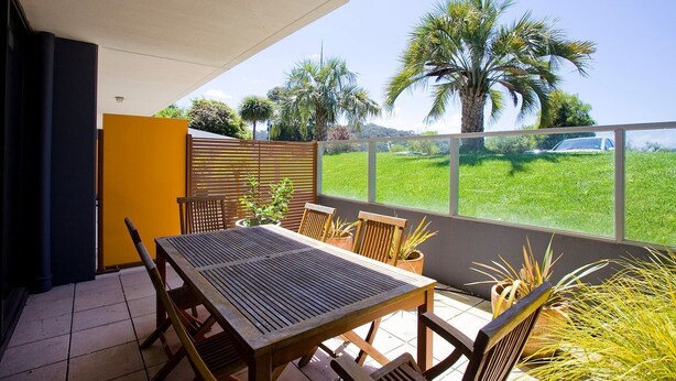 Un patio al aire libre con una mesa y sillas de madera, una pared amarilla y vistas a un césped verde y palmeras.