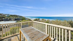 A wooden deck with a table and chairs overlooking a coastal road and ocean. - Beach View (Fairhaven)