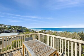 A wooden deck with a table and chairs overlooking a coastal road and ocean. - Beach View (Fairhaven)