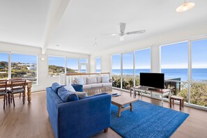 A spacious living room with a large blue sofa, a coffee table, and a TV on a stand. There are large windows offering a view of the ocean and hills.