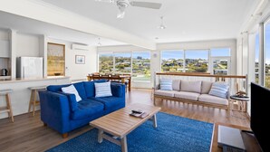 A spacious living area with a blue sofa, a beige sofa, and a wooden coffee table. There is a ceiling fan, a dining table with chairs, and a large window offering a view of the landscape.