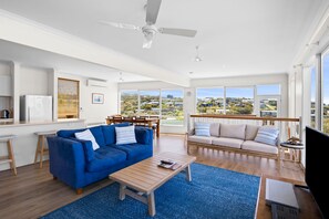 Een ruime woonkamer met een blauwe bank, een beige bank en een houten salontafel. Er is een plafondventilator, een eettafel met stoelen en een groot raam met uitzicht op het landschap.