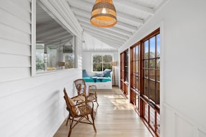 A covered porch with a wooden floor, wicker chairs, a hanging light fixture, and a view of the outdoors through large windows.