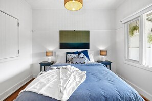 A bedroom with a bed, two bedside tables, a hanging light fixture, a painting on the wall, and a window with a plant. - Sur La Mer (Lorne)