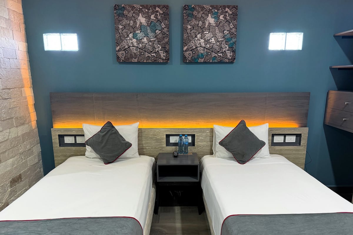 A hotel room with two beds, a nightstand, a wall-mounted TV, and two paintings.
