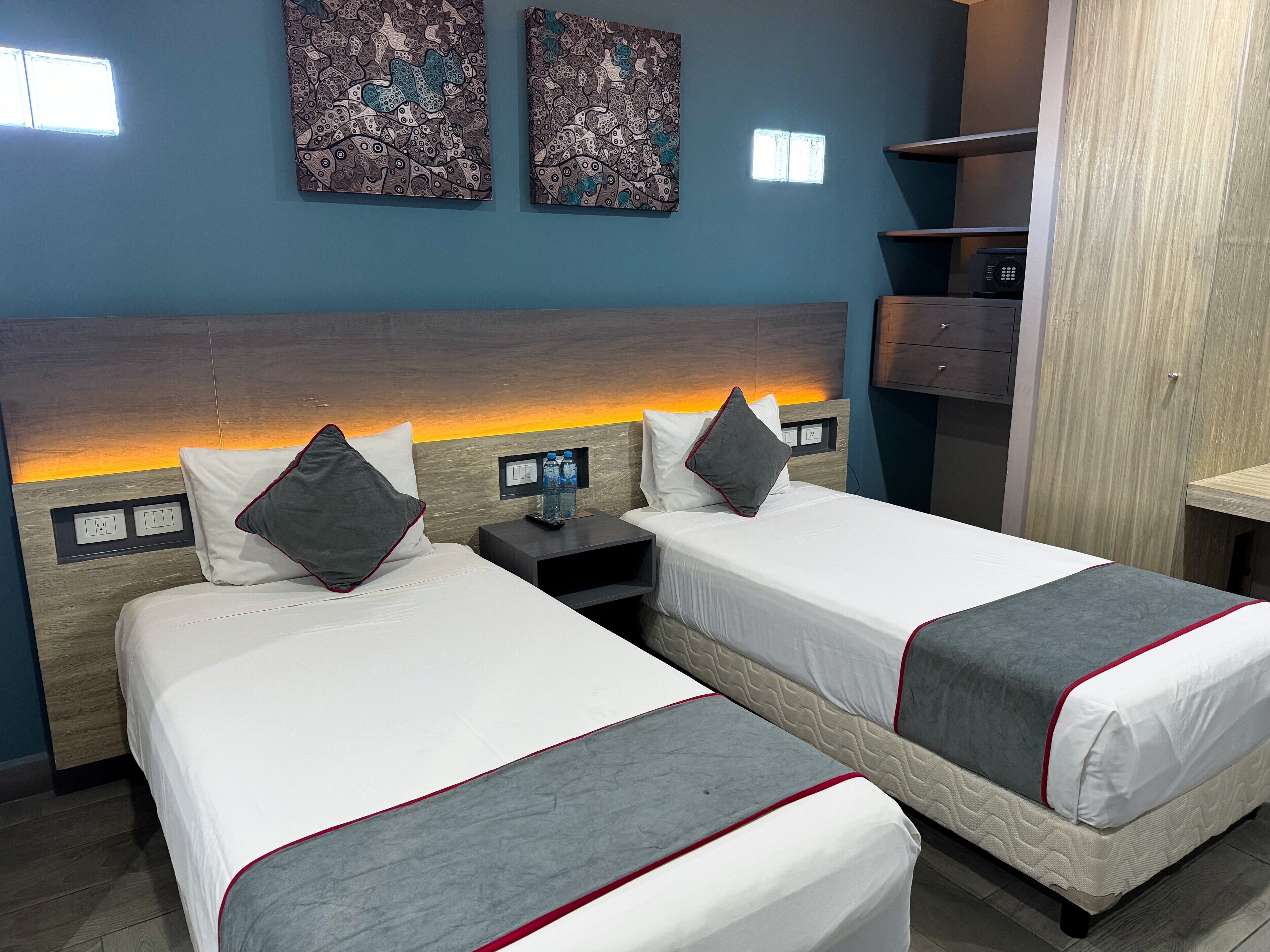 Une chambre d’hôtel avec deux lits, une table de chevet avec une télécommande, une lampe fixée au mur et une étagère avec un téléphone.