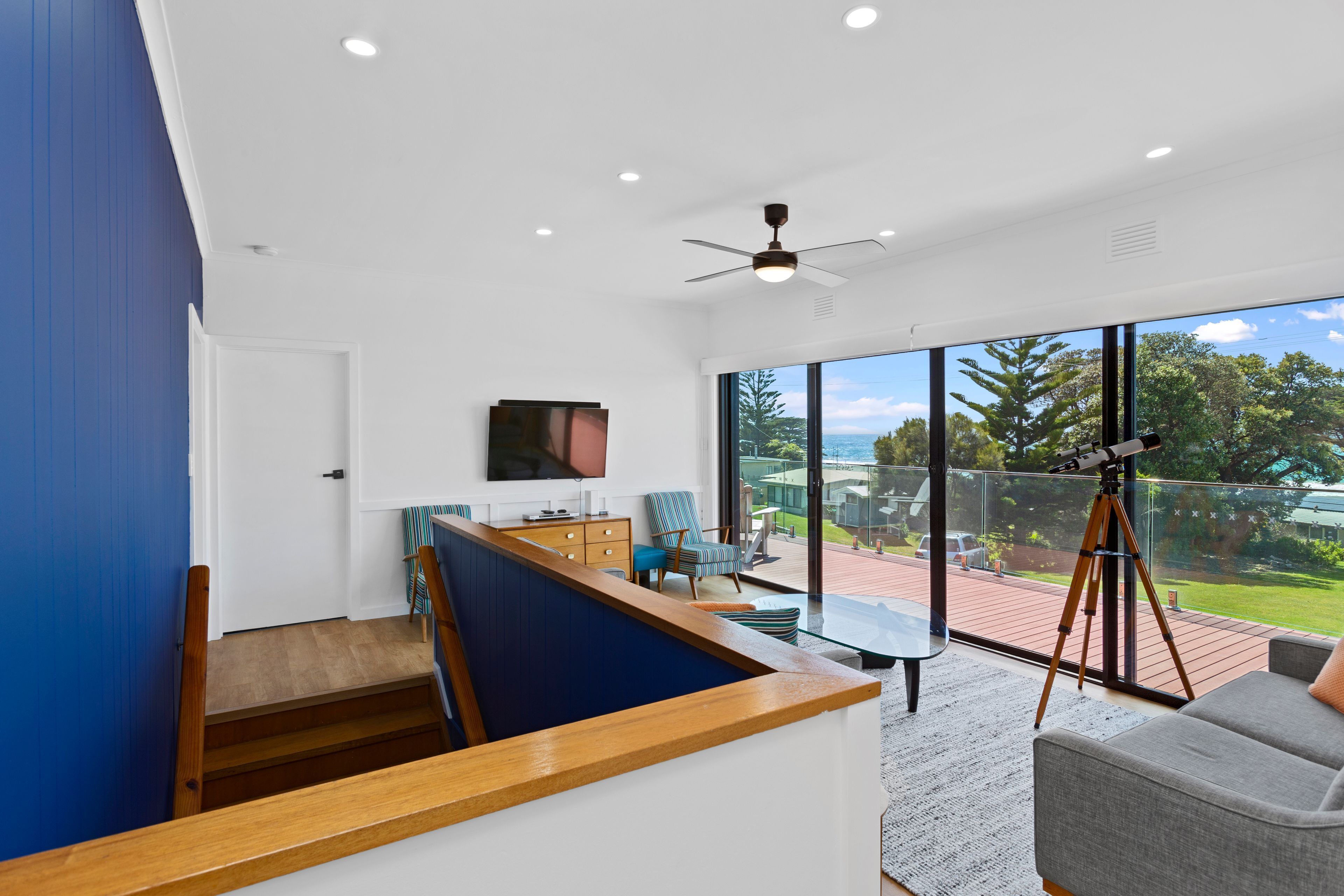 A modern interior with a wooden staircase, a flat-screen TV, a dining area, and a balcony with a telescope.