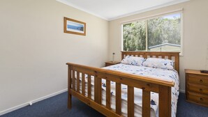 A bedroom with a wooden bed, a nightstand, a dresser, a framed picture on the wall, and a window with a view of trees. - Summer Breeze (Apollo Bay)