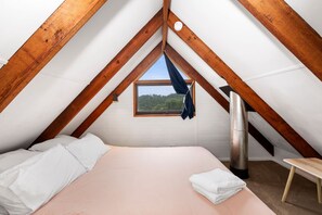 Un dormitorio con techo inclinado, una cama con ropa blanca, una ventana con vista a follaje y un calentador.