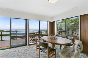 Een ruime kamer met een grote tafel en stoelen, grote ramen met uitzicht op zee, en een balkon met een leuning.