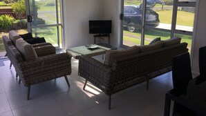 A modern living room with wicker furniture, a flat-screen TV, and large sliding glass doors offering a view of a parking lot and greenery. - stunning 2 bedroom 2 bathroom holiday home with a kiwiana caravan for extra beds (Ohope)