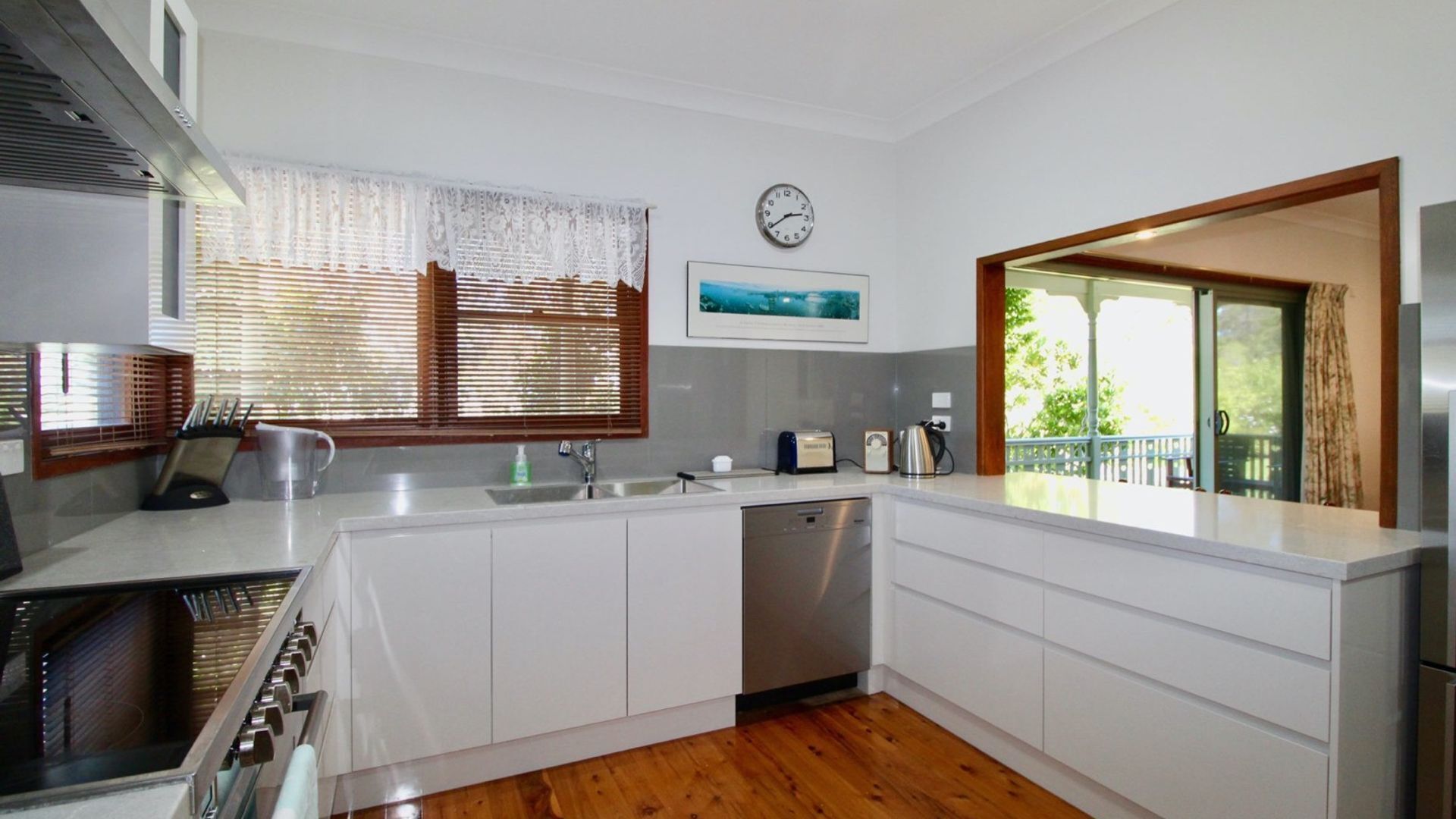 A modern kitchen with white cabinetry, a built-in dishwasher, a stove, and a sink. There is a clock on the wall and a window with blinds.