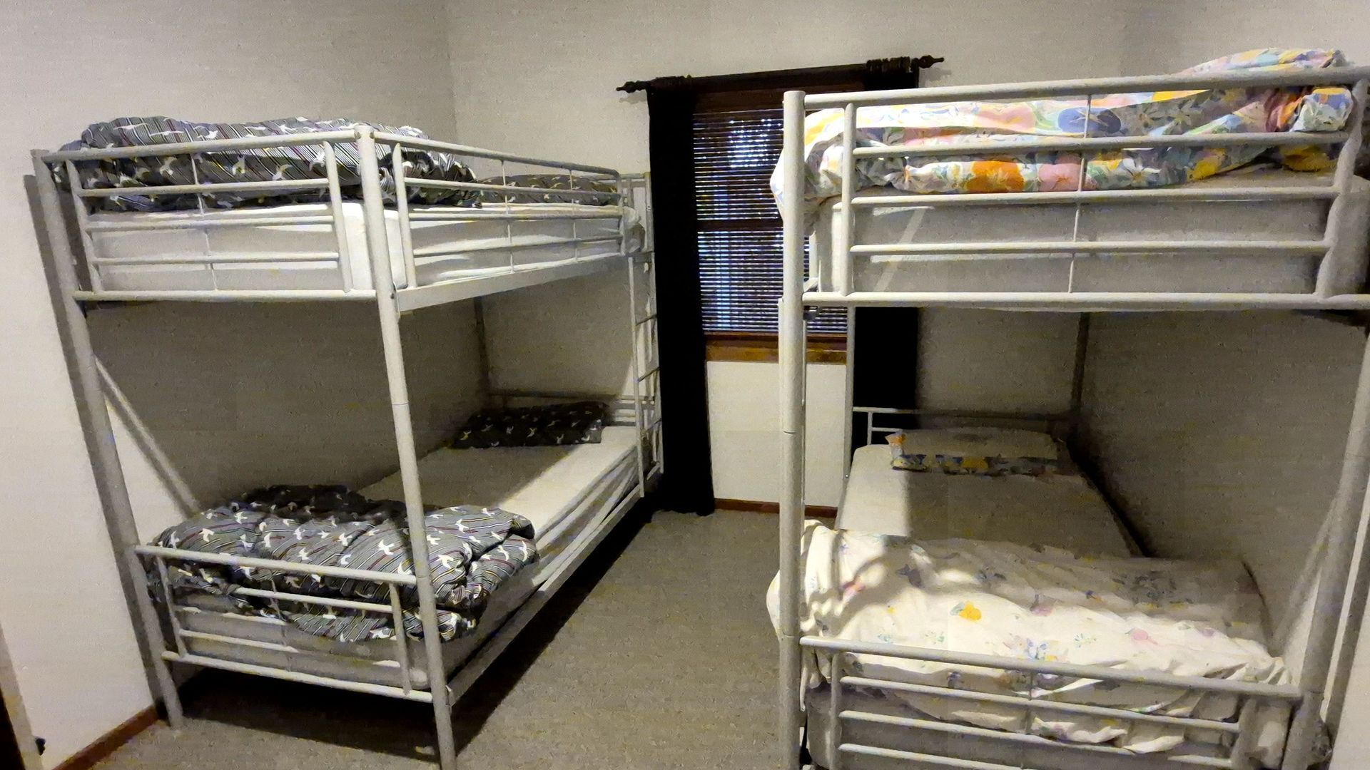 Une chambre avec deux lits superposés, chacun étant équipé d’un matelas et d’une couverture. On aperçoit une fenêtre avec des stores au fond.
