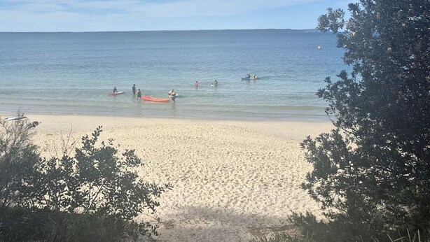 En strand med klart vatten, där folk badar och paddlar, och där det finns båtar.