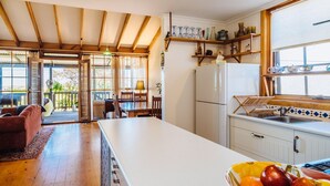 A kitchen with a large island, wooden beams, a dining area with a table and chairs, and a view of the outdoors. - The blue beach house (Quinns Rocks)