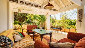 A cozy patio area with a sofa, chairs, and a table, surrounded by lush greenery and a pergola with hanging lights. - The blue beach house (Quinns Rocks)
