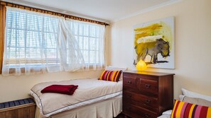 A bedroom with a bed, a wooden dresser, a window with sheer curtains, and a painting on the wall. - The blue beach house (Quinns Rocks)