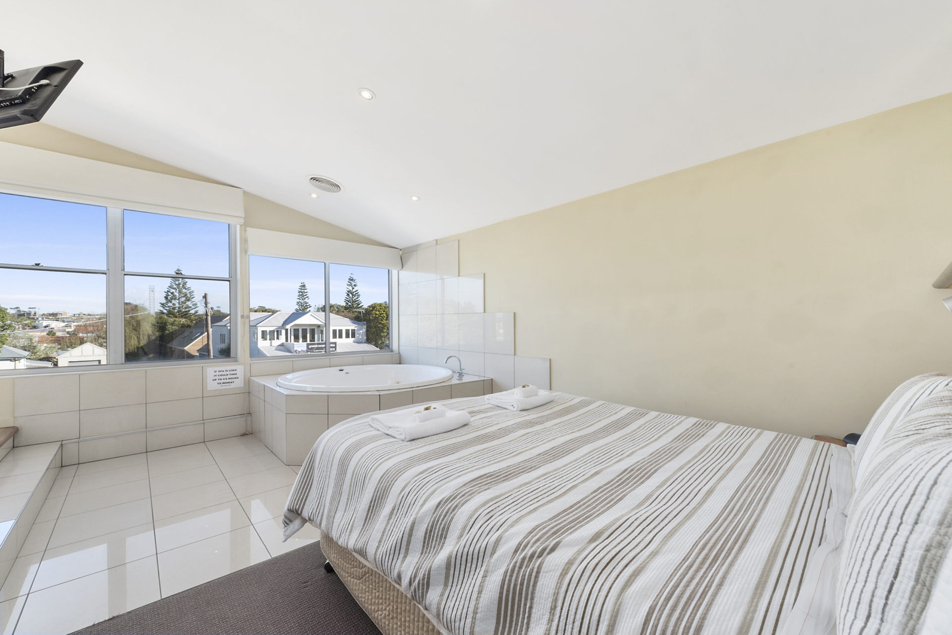 A modern bedroom with a large bed, a bathtub, and a view of a residential area.