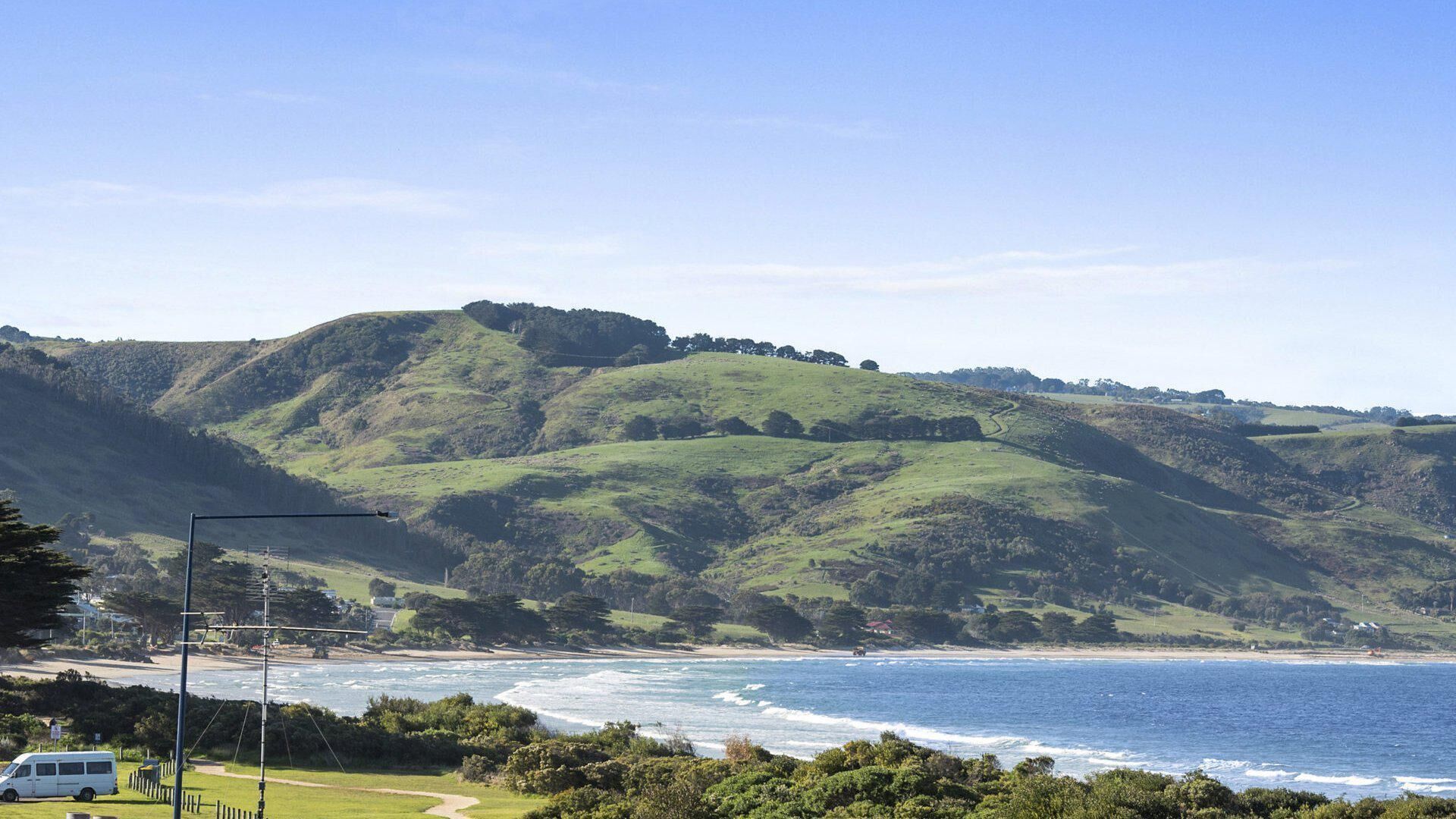 A coastal landscape with rolling hills, a beach, and a white van parked near a pathway.