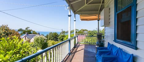 Una terraza de madera con barandilla, un espacio cubierto con ventana y vista al mar y casas al fondo.