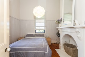 A bedroom with a bed, a hanging light fixture, a window with blinds, a fireplace, and a small table with a vase of flowers. - Valetta (Lorne)