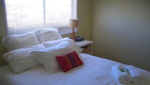 Un lit bien fait, avec des draps blancs, un oreiller rouge et violet, et une serviette enroulée posée sur le côté.