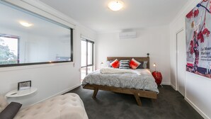 A bedroom with a bed, a bedside table, a bench, a window, and a wall-mounted air conditioning unit. - Allawah (Lorne)