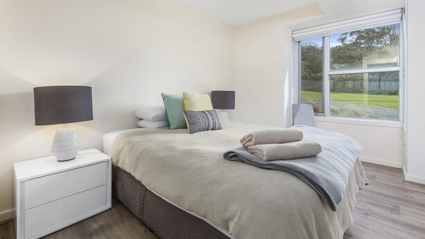 A bedroom with a bed, bedside lamps, a nightstand, a window with a view of the outdoors, and a brick-patterned wall.