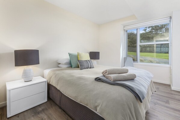 A bedroom with a bed, bedside lamps, a nightstand, a window with a view of the outdoors, and a brick-patterned wall.