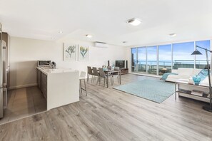 A modern kitchen with a dining area, a living room with a sofa and a view of the sea, and a large window. - Point Grey Apartment 13 (Lorne)