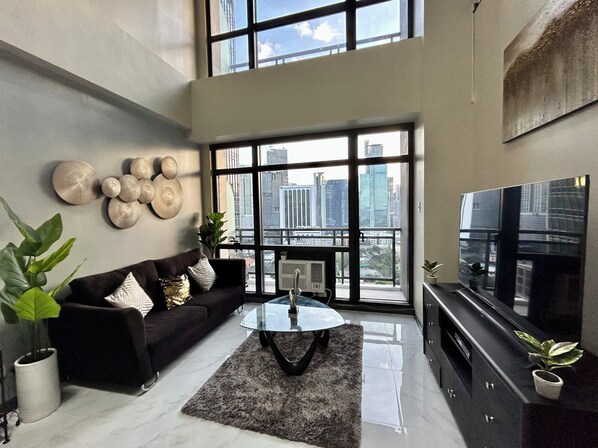 A modern living room with a black sofa, a glass coffee table, and a large TV. There are decorative wall pieces and a view of the city through large windows. - Stunning 2 Floor Loft w/ 5 Balconies & Round Bed (Makati)