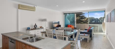 A modern kitchen with a built-in dishwasher, a dining area with a large window, and a living space with a sofa and a TV.