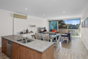 A modern kitchen with a built-in dishwasher, a dining area with a large window, and a living space with a sofa and a TV. - The Foreshore Apartment (Apollo Bay)