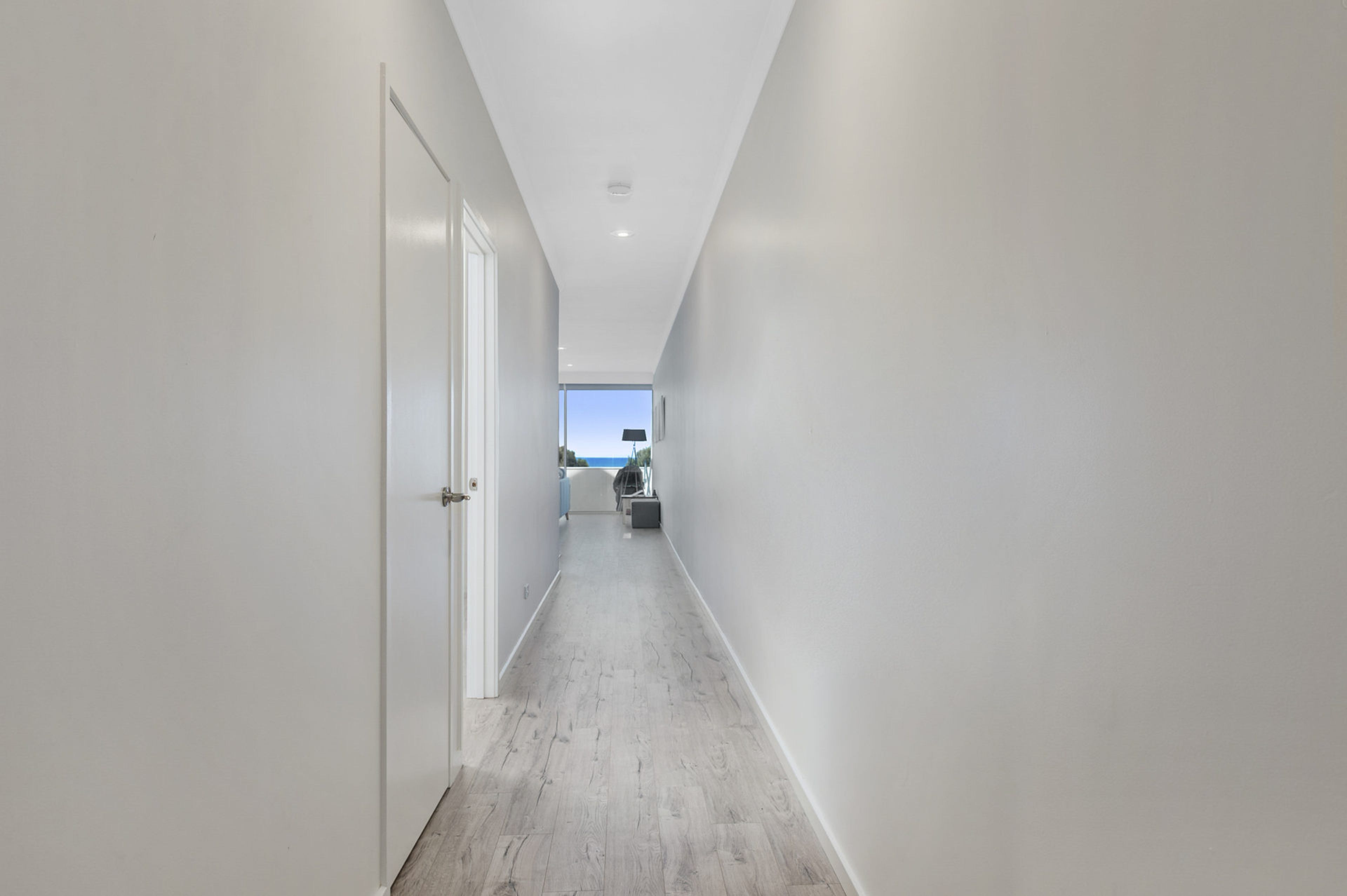 Un couloir étroit aux murs blancs et au plancher en bois, qui mène à une pièce offrant une vue sur le ciel.