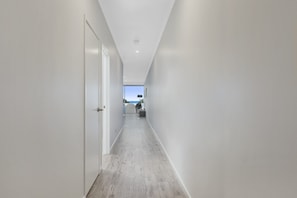 A narrow hallway with white walls and a wooden floor, leading to a room with a view of the sky. - The Foreshore Apartment (Apollo Bay)