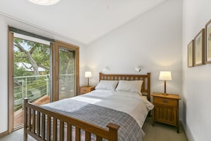 A bedroom with a wooden bed, two bedside tables with lamps, a large window with blinds, and a balcony with a view of trees.
