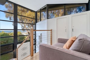 A modern interior with a view of the sea, a staircase with a wooden railing, and a sofa with cushions. - Skenes Beach House Ocean Views set in Natural Bush (Skenes Creek, Apollo Bay)