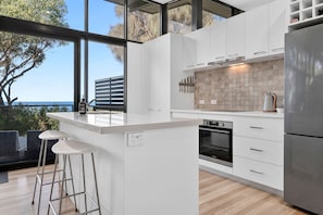 A modern kitchen with white cabinetry, a stainless steel refrigerator, and a large window overlooking a scenic view. - Skenes Beach House Ocean Views set in Natural Bush (Skenes Creek, Apollo Bay)