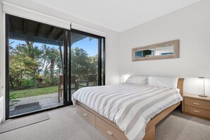 A bedroom with a large sliding glass door leading to an outdoor area, a bed with a striped bedspread, a bedside table with a lamp, and a mirror on the wall. - Skenes Beach House Ocean Views set in Natural Bush (Skenes Creek, Apollo Bay)