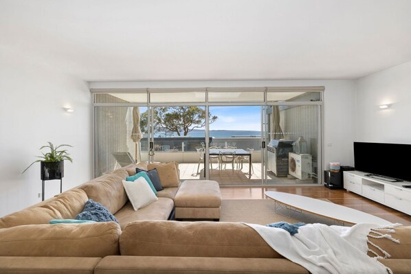 A modern living room with a large sofa, a flat-screen TV, and a balcony with a table and chairs.